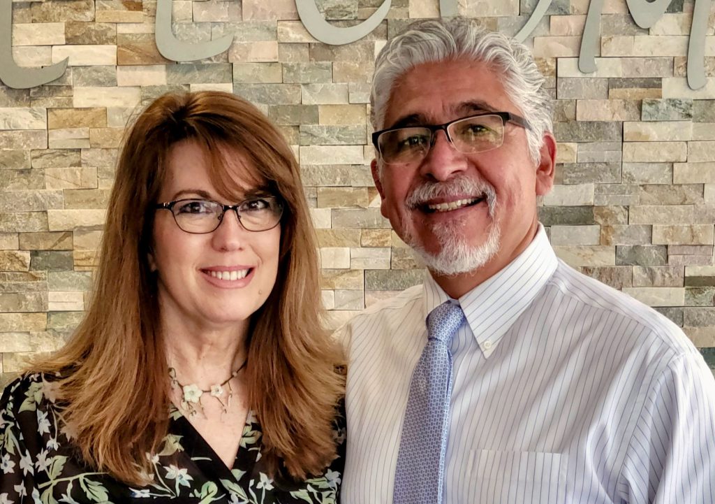 A woman with shoulder-length auburn hair and glasses and a man with gray hair and glasses wearing a dress shirt and tie stand close together smiling in front of a stacked-stone accent wall.