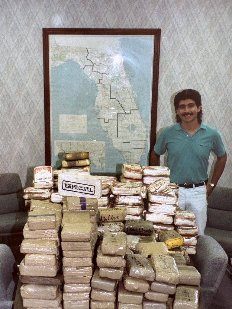 A man in a teal polo shirt stands smiling behind a large table stacked with wrapped drug bricks, with a framed Florida map on the wall behind him