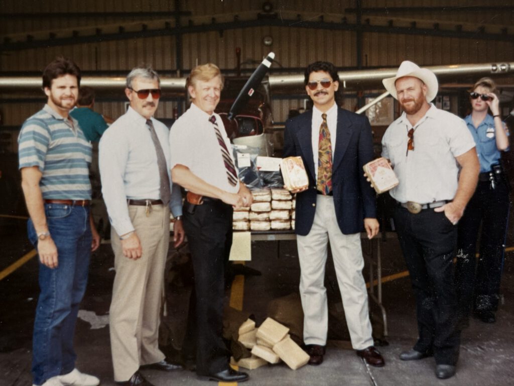 Five men stand in an aircraft hangar in front of a small plane and a table stacked with wrapped drug bricks; two men in the center shake hands while others hold individual bricks