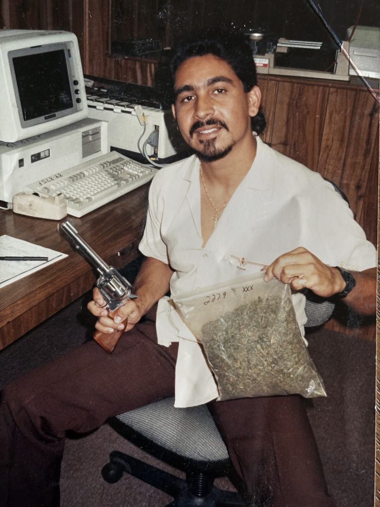 A bearded man seated at a desk holds up a labeled evidence bag and a revolver, with an early computer terminal visible to his left