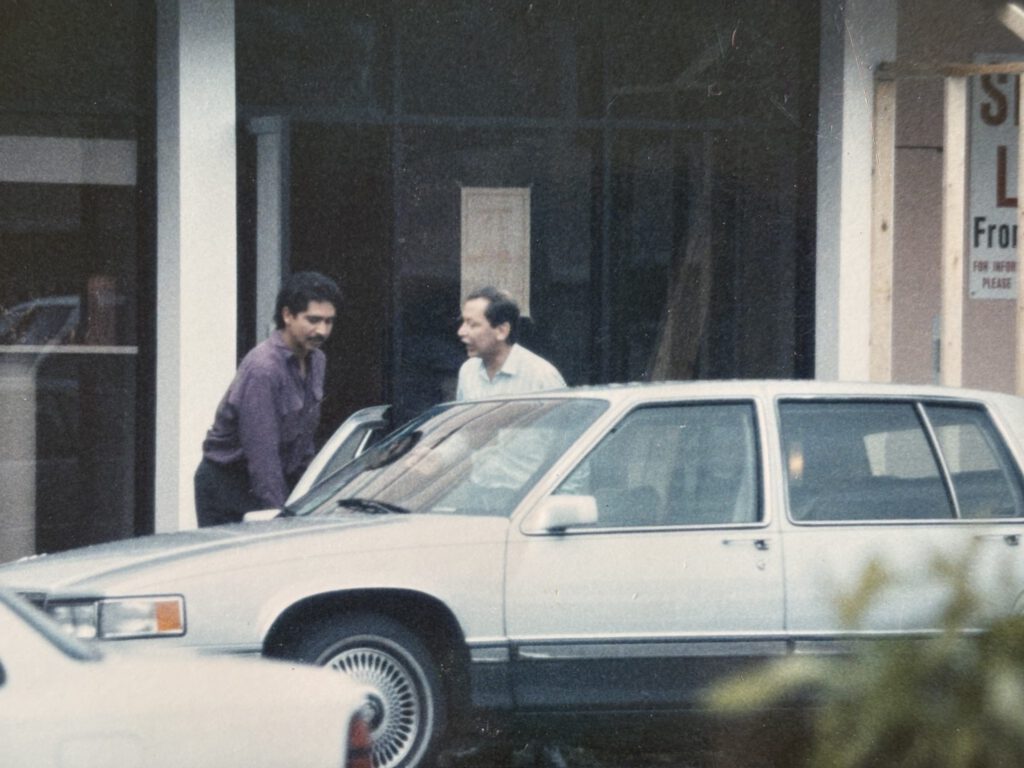 Grainy surveillance-style photograph of two men standing near a white van outside a building, shot from a distance at an angle