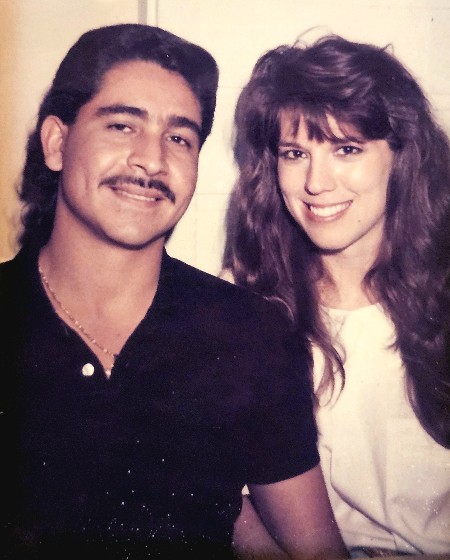 A man with a mustache and a woman with long wavy hair pose together smiling in a close portrait, vintage early-1990s photograph