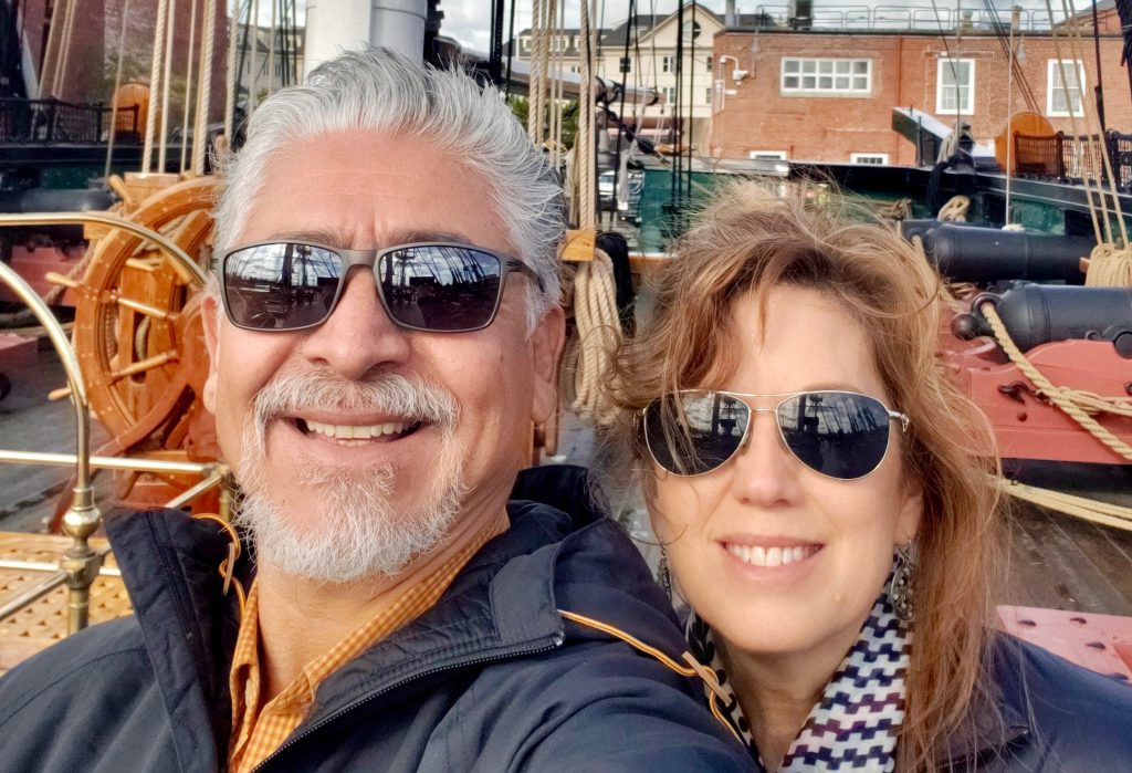 A silver-haired man with a white beard and sunglasses and a woman with auburn hair and sunglasses smile together in a selfie taken on the deck of a tall ship at a historic waterfront, rigging and brick buildings visible behind them.